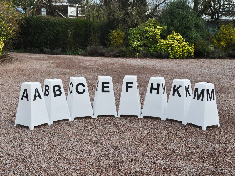 8 Dressage Tower Markers sat on gravel. Black painted letters on all sides (A, B, C, E, F, H, K, M).