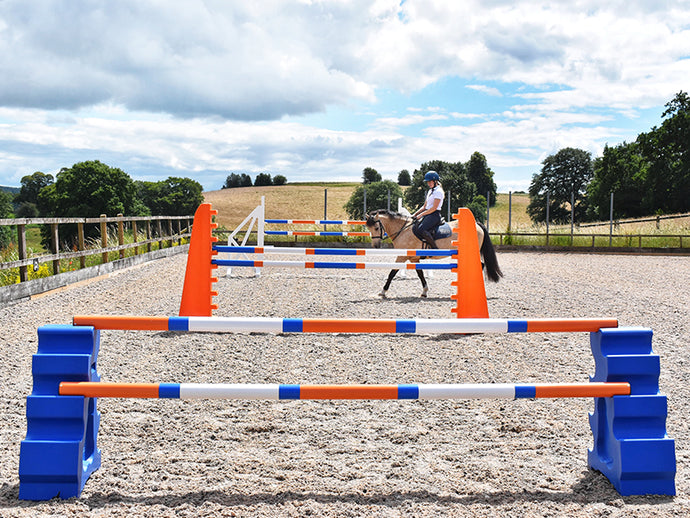 Blue MultiJumps with 9 Band Poles coloured Orange, Blue and White. Behind that, 2 orange 8 Cups with 9 band poles coloured: Blue, Orange and white. At the back White Cross wings with 9 band poles coloured Blue, white and orange. All jumps in arena with horse and rider walking between jump sets. 